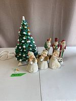 Full view of vintage ceramic lighted Christmas tree with three angel and three Santa figurines arranged beside it on a table with a neutral curtain background.