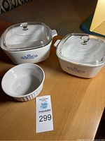 Two square white Corningware casserole dishes with blue floral tulip pattern and clear glass lids, alongside a white round 16 oz stoneware dish on wooden surface.