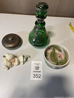 Four vintage items on white table: green glass decanter with stopper, round pottery container with floral design, Lenox sleeping child figurine, and small metal case.