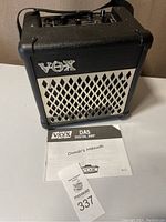 Front view of black VOX DA5 digital amplifier with white grille and VOX logo, placed on white surface together with owner's manual.