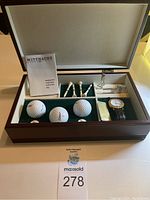 Overview of the open wooden box with contents - golf balls, tees, watch, divot tool, gold clip, and warranty booklet visible