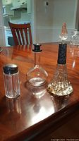 Three glass condiment servers on wooden table: salt shaker, oil bottle, and tall vinegar bottle with decorative sterling silver parts.