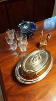 Overall view of six liqueur glasses, blue carnival glass dish, condiment fork set with wooden holder, and silver chafing dish on wood surface.