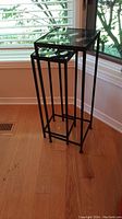 Two metal nesting tables with glass tops, arranged nested near a window on a wood floor. Both tables have thin black metal frames and clear glass tops.