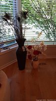 Tall black floor vase with faux peacock feathers and smaller ceramic vase with faux white orchids and red roses, placed on wooden floor in front of window with blinds.