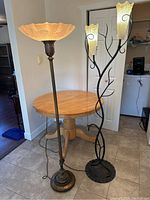 Photo showing two standing lamps, one with a large round floral glass shade and one with three yellow flower-shaped shades on black vine-like metal base, positioned near a wooden table.