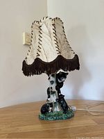 Photo of a ceramic lamp with a tree trunk base and three climbing black bears, topped with a cream-colored lampshade with dark brown fringe.