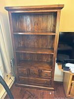 Full view of the wooden bookshelf showing three shelves and four drawers, placed against wall and next to a TV stand.
