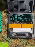 Open hard plastic carrying case containing a green hand drill press with accessories and manuals inside.