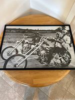 Black and white professionally framed Easy Rider movie poster showing two motorcycles riders shaking hands, one on a chopper with American flag design. Framed in black wood frame on round wooden table.