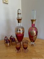 Photo showing two ornate glass decanters with stoppers and six accompanying red glass drinking glasses with gold accents, displayed on wooden surface against white wall.