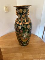 Front view of tall green vase with gold ornate handles and detailed floral designs on body, placed on wooden table.