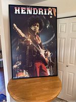 Front angled view of the large framed Jimmy Hendrix poster showing the entire frame and part of the wooden table it is resting on.