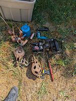 Photo showing miscellaneous tools - including vintage oil cans, a plastic container, two metal pulleys, a power drill with cord and battery, a red clamp, and various grease tools scattered on grass.