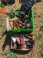 Top-down photo showing a green plastic crate filled with assorted tools including wooden and metal hand tools, a fishing reel, metal clamp, and two vintage license plates. A cardboard box in the foreground contains sanding belt on spool, wire strippers, tie wire, and other small hardware items.