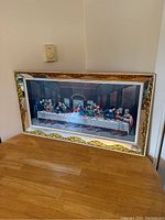 Framed print of The Last Supper resting on a wooden surface against a wall, showing full view of art and frame.