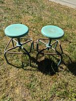 Two vintage-style metal shop stools with turquoise rotating seats and black metal frames placed on grass outdoors.