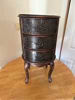 Front view of the small round side table showing three drawers with ring pulls and detailed carved tropical patterns.