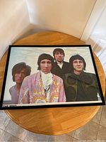 Framed poster of The Who band in colorful 1960s clothing, looking at camera from waist up, sitting on round wooden table, shown flat from above