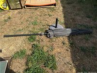 Outdoor patio umbrella folded on the ground with metal pole and attached fabric canopy, mostly gray color, with umbrella stand visible underneath.
