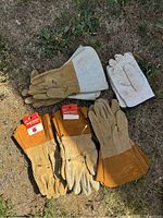 Five pairs of welding gloves positioned on grass showing two pairs without brand tags and three Watson brand tagged pairs mainly in tan and orange suede or leather.