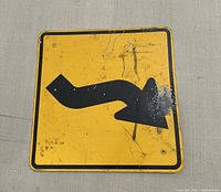 Front view of square metal road sign with double curve arrow symbol, showing wear and scratches.