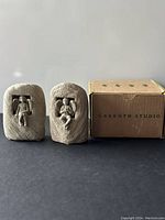 Front view of two cast stone angel figurines with original box. Each figurine depicts an angel seated in a niche within a stone-like textured rectangular block.
