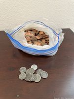 Plastic bag filled with copper pennies with a smaller collection of steel pennies displayed outside the bag on a dark wooden surface.