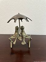 Front view of the brass frog sculpture showing the two frogs seated on a bench under the umbrella