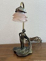 Full view of the vintage Art Nouveau table lamp showing bronze-tone metal base with female figure and pink frosted glass shade.