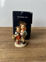 Front view of vintage M.I. Hummel boy and girl porcelain figurine with original box behind it.
