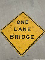 Full view of the vintage metal 'ONE LANE BRIDGE' sign showing its diamond shape, yellow background, black text and border, with visible signs of wear.