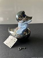 Front and side views of the vintage plated silver Danish duck-shaped bank, showing the blue glass eyes, knitted blue scarf, and accompanying metal key on a dark surface.