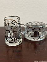 Photo showing the clear glass creamer and sugar bowl with sterling silver overlay designs on a wooden surface against a plain background.