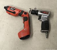 Two tools laid on a surface: an orange and black cordless screwdriver and a pneumatic air hammer.