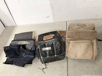 Three tool pouches side by side on a floor: gray and blue fabric pouch, black leather pouch, tan leather pouch.