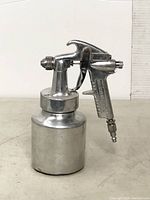 Front perspective view of the Campbell Hausfeld metal spray gun with attached container on a flat surface.