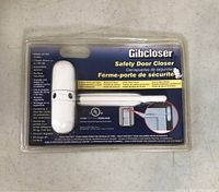 Front view of the Gibcloser safety door closer in clear plastic packaging showing white plastic device and printed cardboard backing with branding and basic usage illustration.