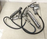 Five assorted air tools laid out on the floor including air ratchet, die grinder, impact wrench, grease gun, and paint spray gun, all with attached hoses, showing wear.