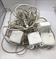 Lot of five white Apple MacBook charger adapters tangled together in a white background, showing varying wear and some dirt marks.