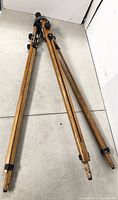 Full length view of the wooden tripod folded on the floor showing three wooden legs with metal fittings at top and plastic/reinforced tips at bottom.