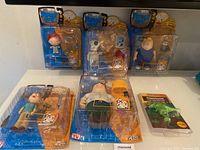 Five unopened Family Guy character figurines in original packaging including Stewie, Brian, Chris, Lois, and Peter, plus a dinosaur candle holder figurine in blister package.