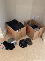 Two medium-sized cardboard boxes each filled with kids winter clothing items including pants and jackets. Various hats and gloves placed in front of boxes on carpet.