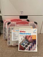 Two plastic cases with red handles containing labeled electronic components alongside a paperback book titled 'Make: Electronics' by Charles Platt