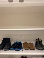 Four pairs of men's shoes displayed on a white shelf: black rubber boots, black and blue Nike sneakers, brown slippers, and gray suede boots.