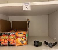 Shelf displaying a cardboard box containing several packs of toe warmers alongside two men's wristwatches, one with a digital display and one analog.