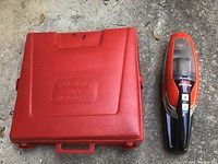 Closed view of the red Ride-A-Way auto emergency kit hard plastic case next to a black and red handheld vacuum cleaner.