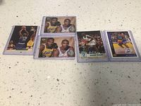 Five basketball cards displayed on a surface, with two in protective sleeves featuring players in Lakers and Minnesota Timberwolves uniforms.