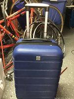 Blue small hard body luggage with visible retractable handle and wheels placed near bicycles.