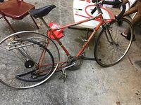 Side view of vintage Crescent 10 speed racing bicycle with orange frame, black saddle, rear metal rack, drop handlebars, missing tires, showing signs of use and minor rust.